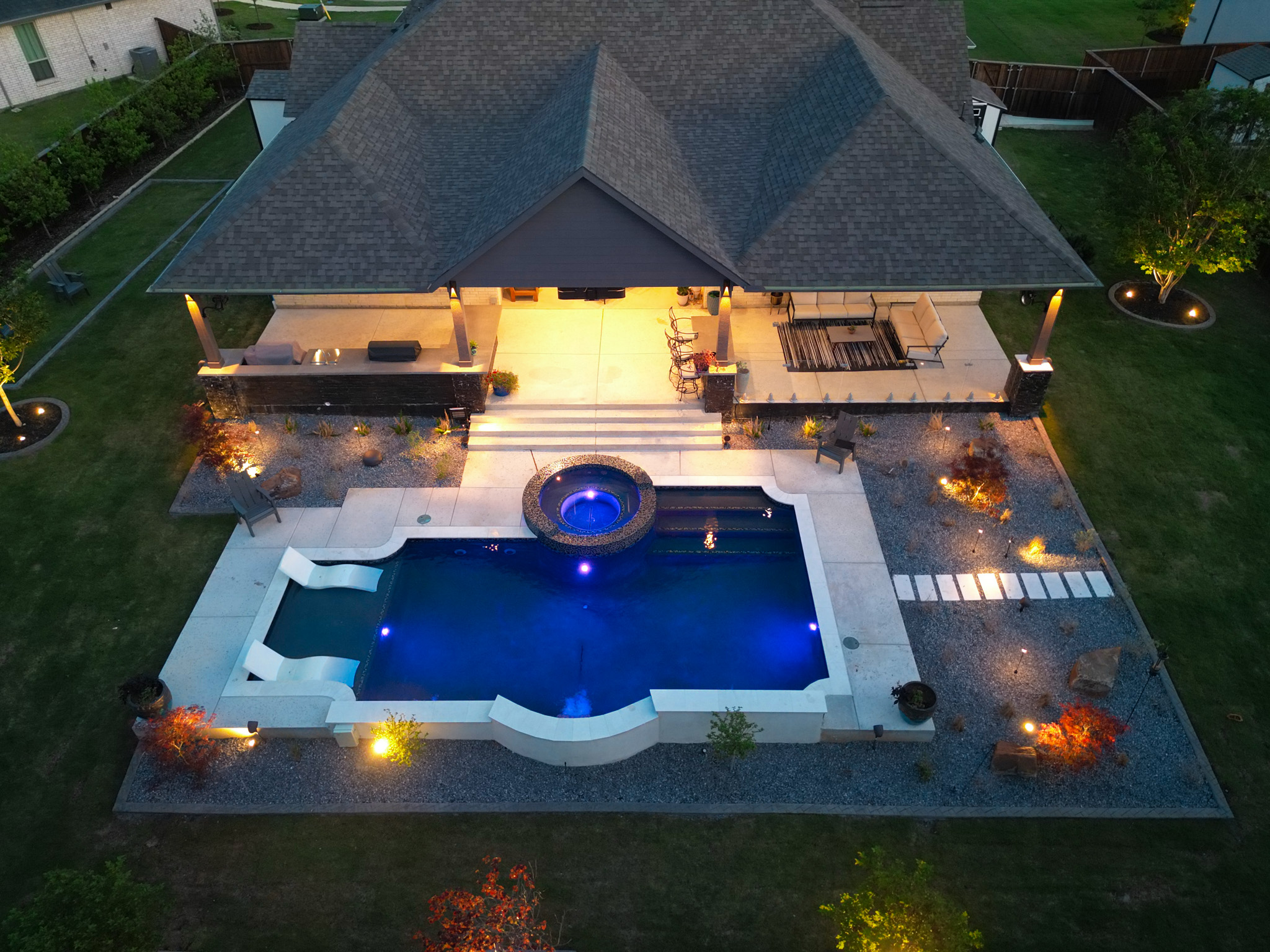 Luxury pool and spa design featuring water features, lounge chairs, and landscape lighting at dusk.